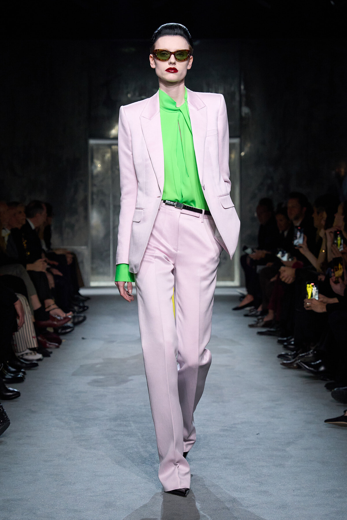 Model on a runway wearing a pastel pink suit with a neon green blouse and dark sunglasses, red lipstick.