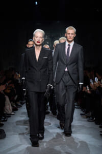 Two models walk a fashion runway in formal suits, with a dark audience on both sides of the catwalk.