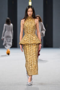 Female model walks a runway wearing a gold textured, high-neck peplum dress with a long straight skirt and metallic blocks, paired with light gray shoes.