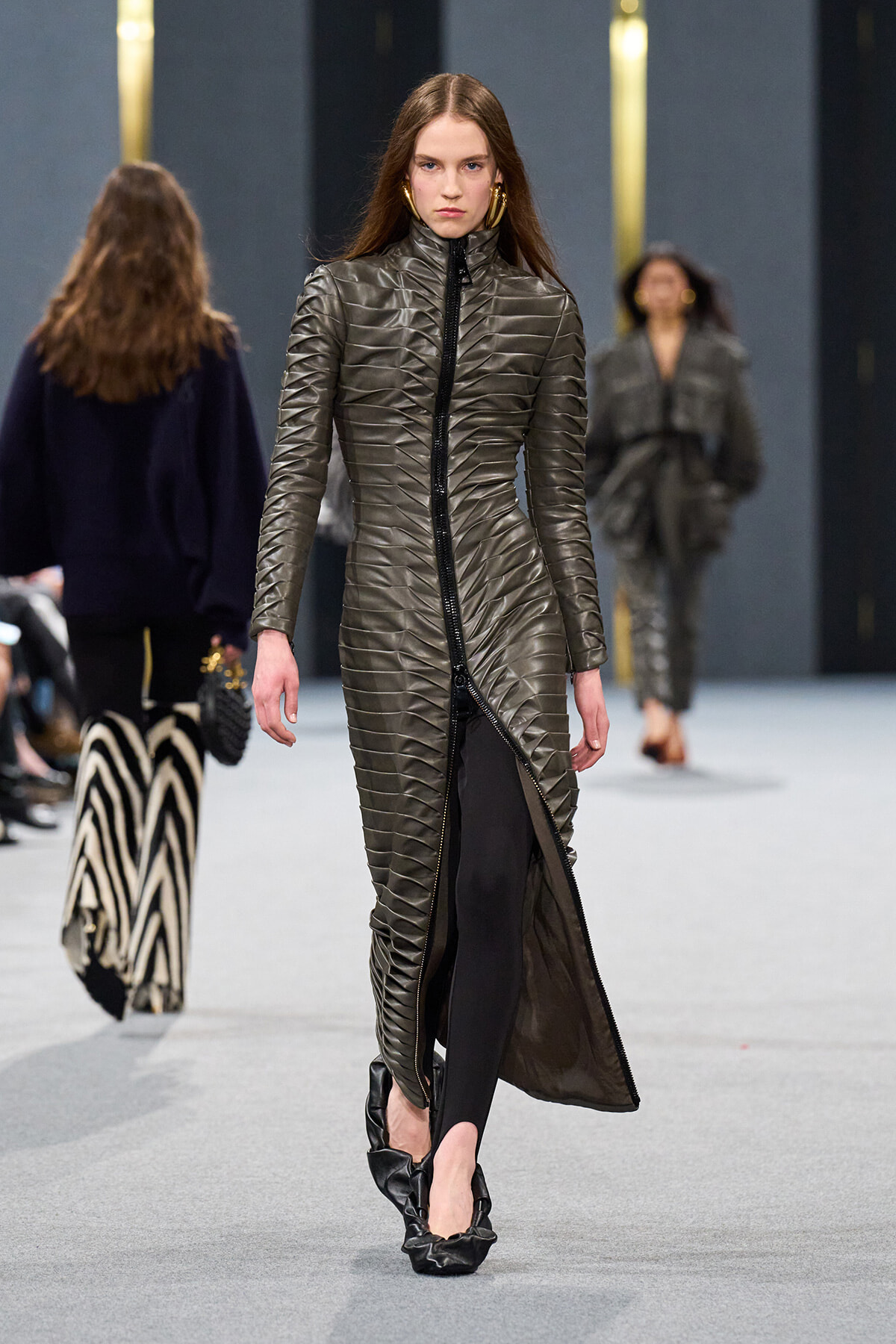 Model walking a fashion show in a long, metallic quilted coat with a high collar and oversized zip, paired with black leggings and black ruffled flats.