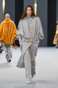 Model on a runway wearing a loose gray wrap coat with a wide belt and matching gray pants; gold buckle detail.