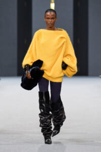 Model walking a runway in a bright oversized yellow knit sweater, dark tights, and slouchy black knee-high boots, holding a dark fur clutch across her body.