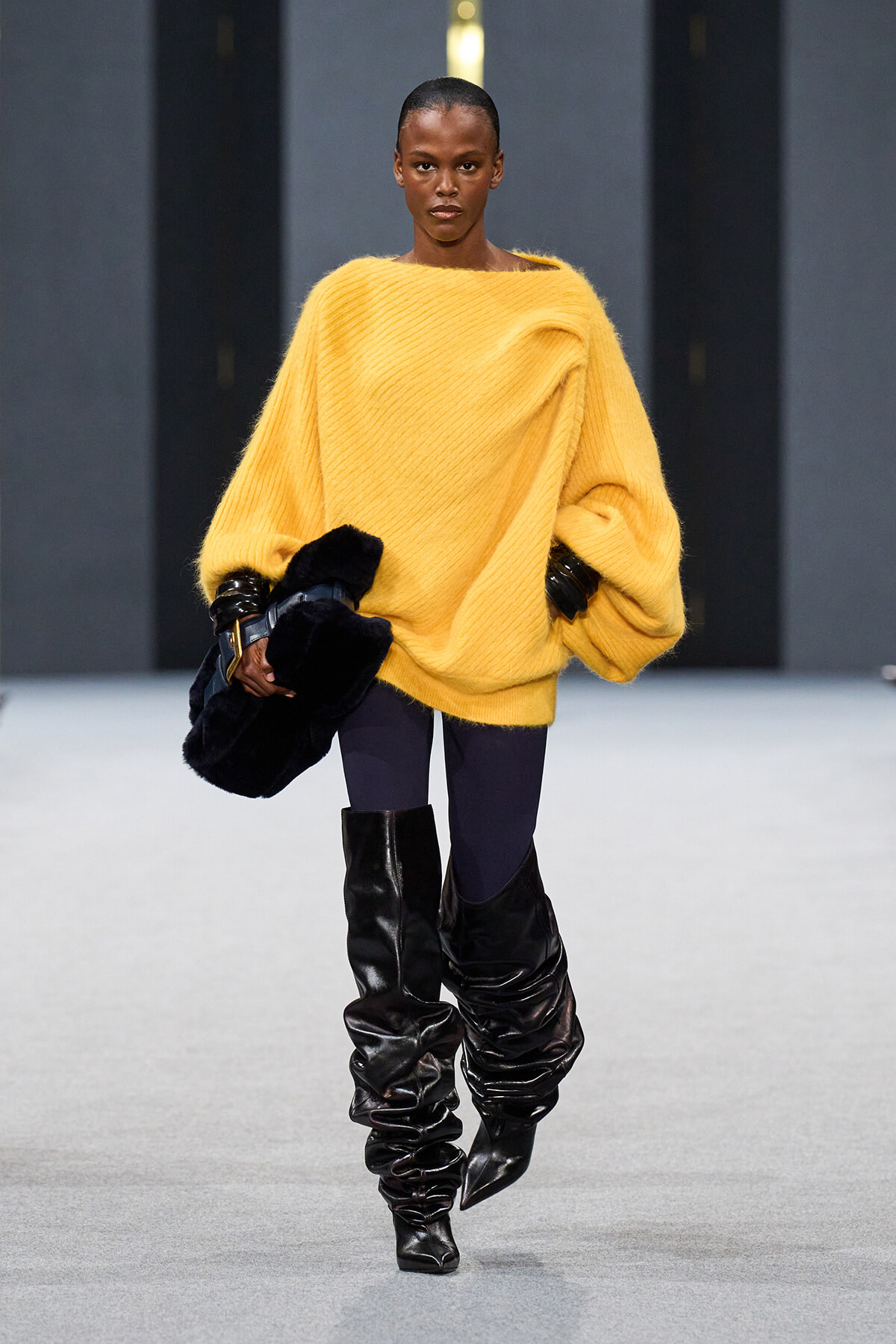 Model walking a runway in a bright oversized yellow knit sweater, dark tights, and slouchy black knee-high boots, holding a dark fur clutch across her body.
