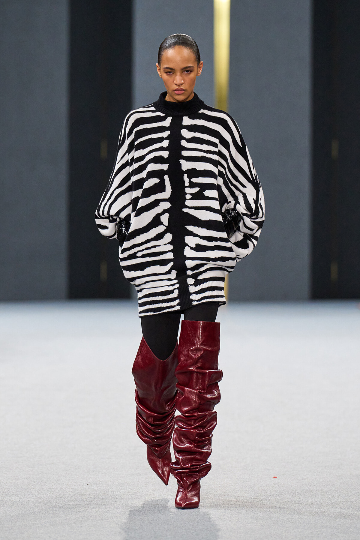 Model walking runway in a black-and-white zebra-striped oversized sweater with black accents and bright red slouchy knee-high boots.