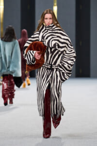 Model walking a runway in a black-and-white zebra-print oversized coat, belted at the waist, clutching a brown fur stole and wearing burgundy knee-high boots on a gray runway.