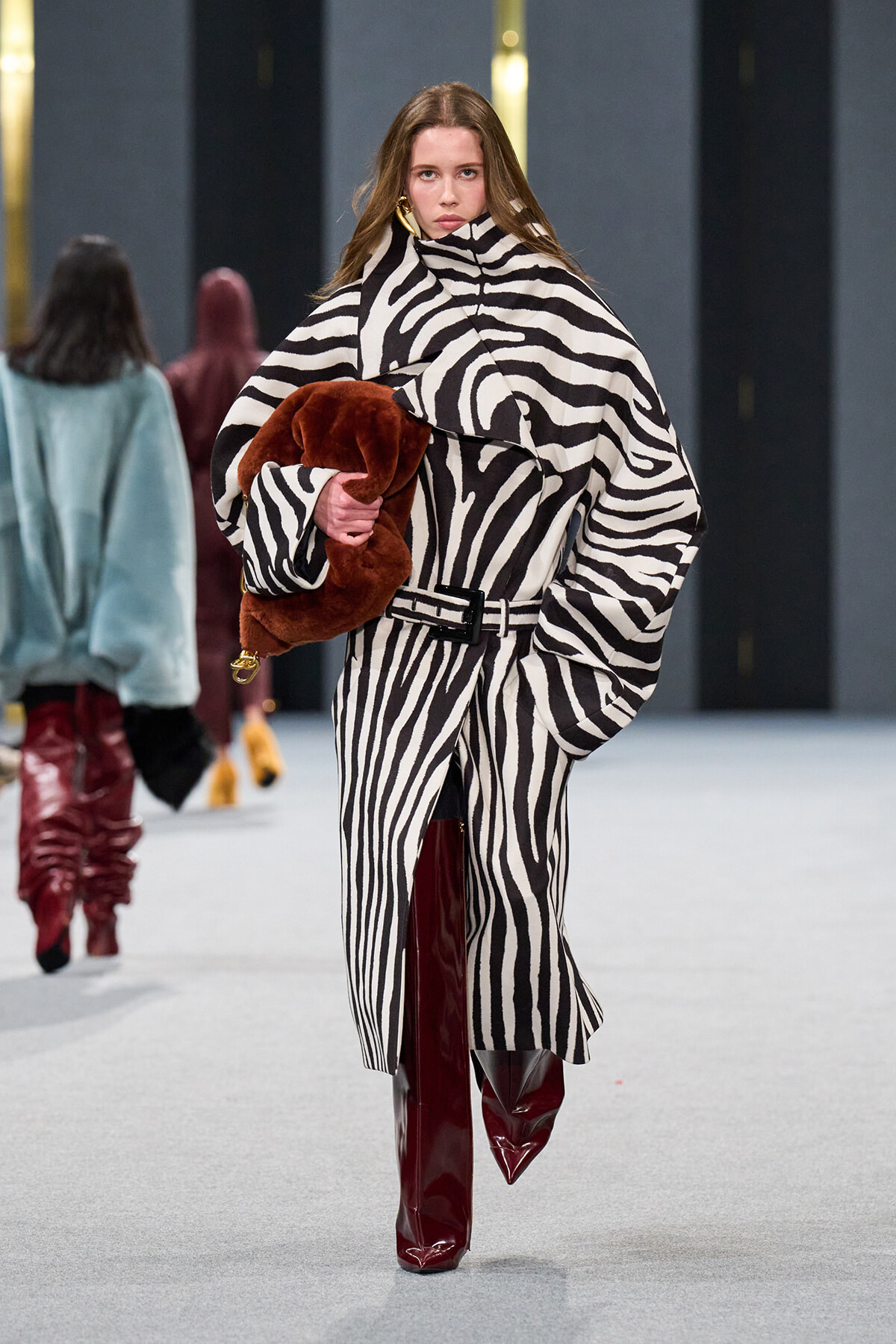 Model walking a runway in a black-and-white zebra-print oversized coat, belted at the waist, clutching a brown fur stole and wearing burgundy knee-high boots on a gray runway.