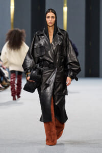 Model walking a runway in an oversized black leather trench with hood, belt, and large pockets, paired with rust-colored furry pants and a black handbag.