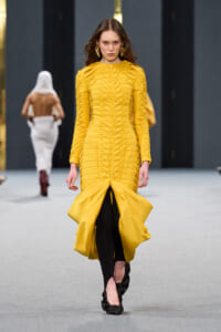 Model walks a runway in a sculptural yellow midi dress with ruching and a dramatic flared hem, wearing black leggings underneath and large gold earrings