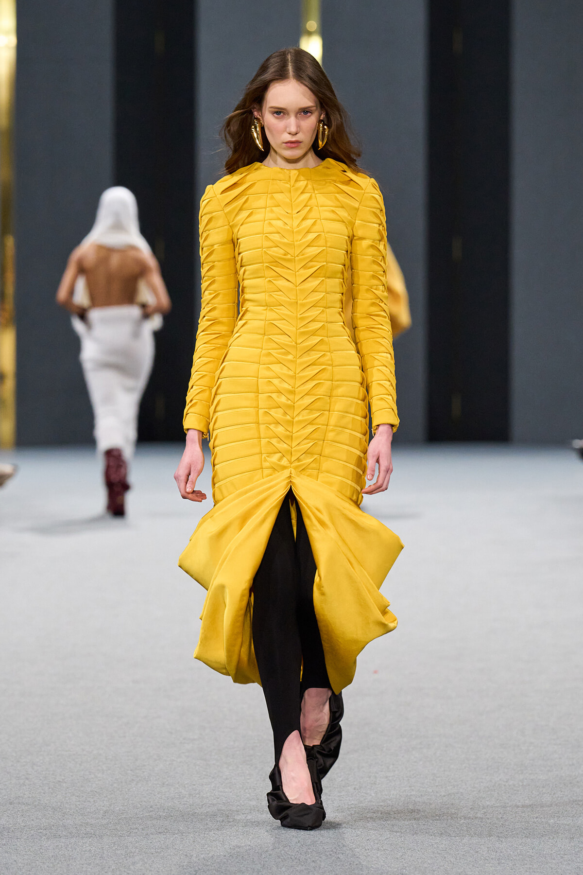 Model walks a runway in a sculptural yellow midi dress with ruching and a dramatic flared hem, wearing black leggings underneath and large gold earrings