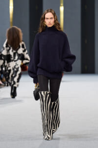 Model walks the runway in a navy oversized knit turtleneck sweater, black leggings, and bold zebra-print knee-high boots, carrying a small bag.