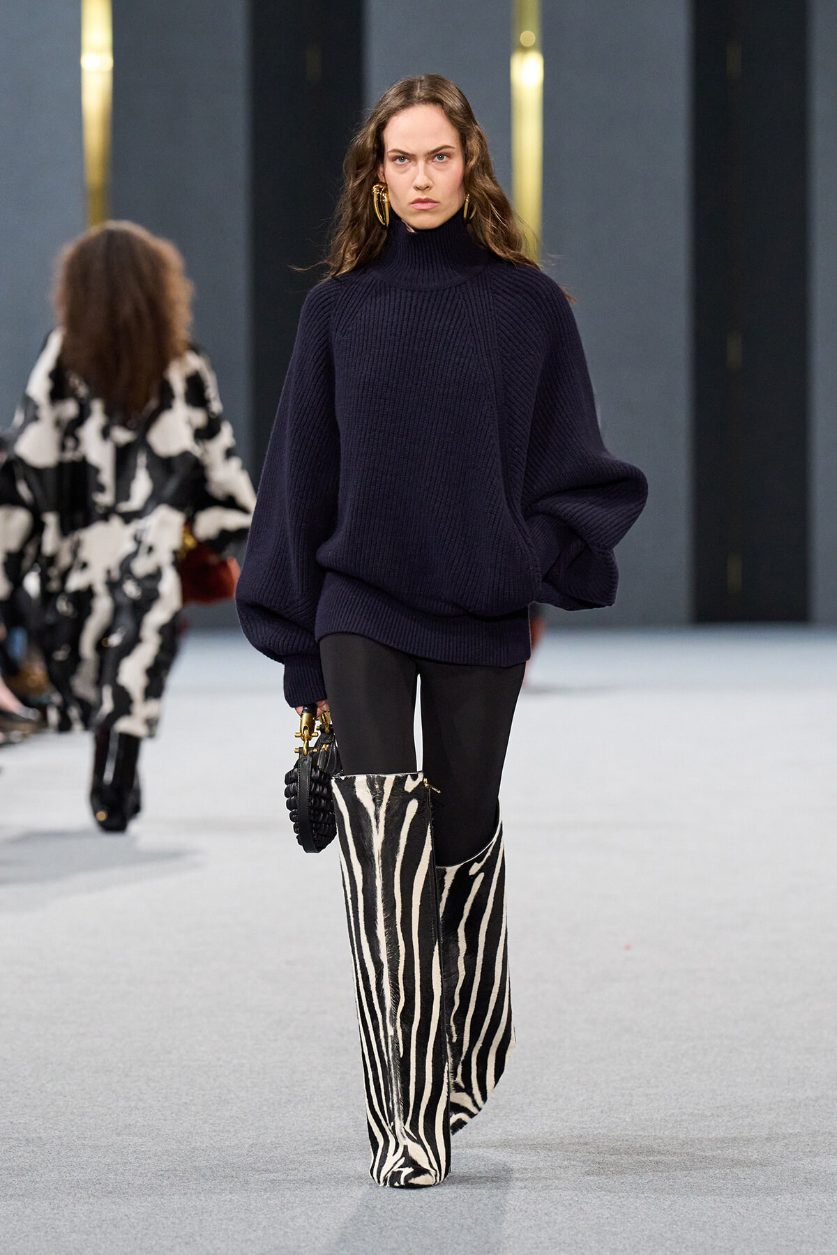 Model walks the runway in a navy oversized knit turtleneck sweater, black leggings, and bold zebra-print knee-high boots, carrying a small bag.