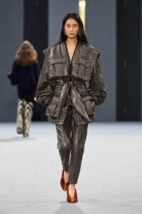 Model on a runway wearing a metallic gray quilted oversized jacket with a tied waist and matching pants, brown suede heels.