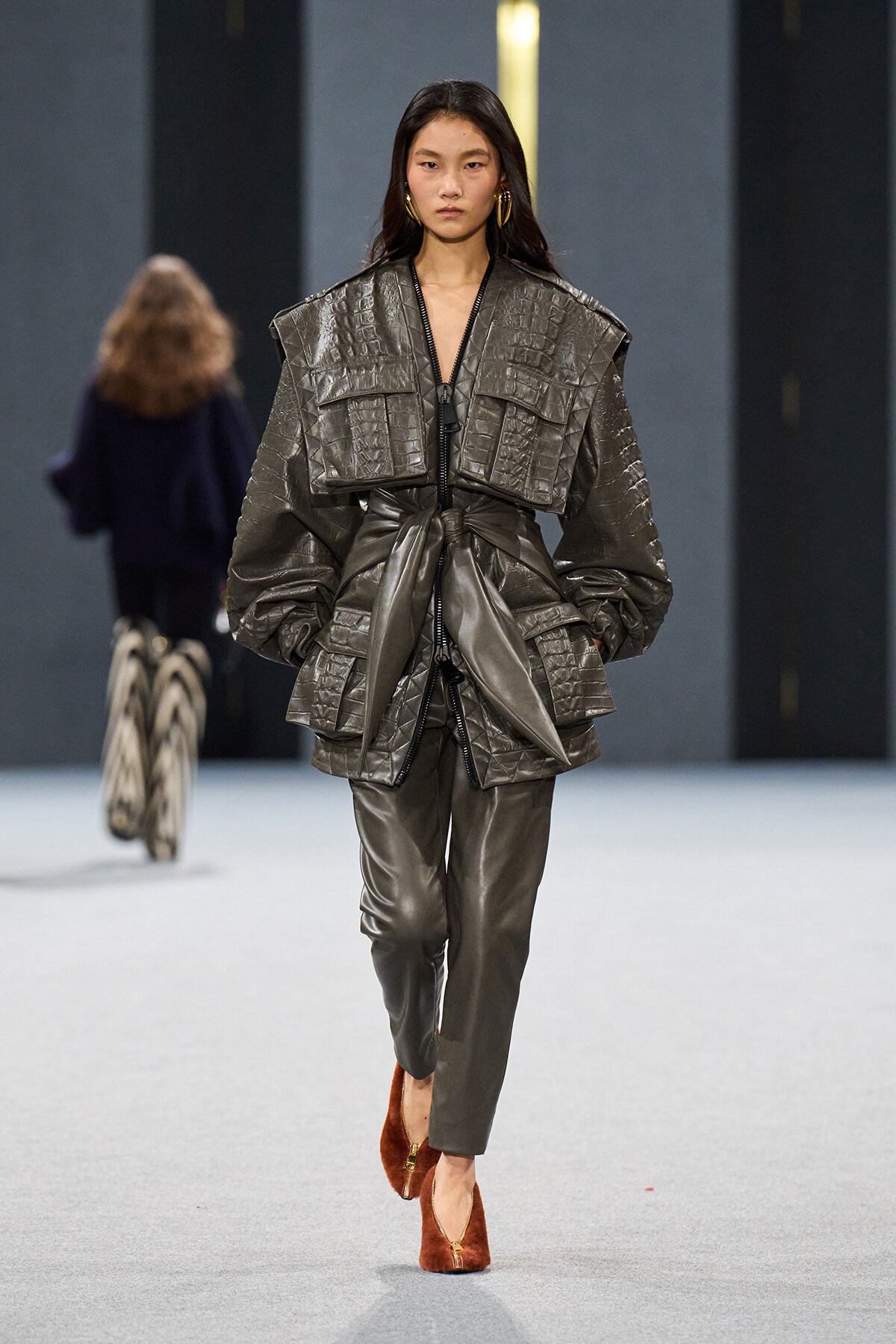 Model on a runway wearing a metallic gray quilted oversized jacket with a tied waist and matching pants, brown suede heels.