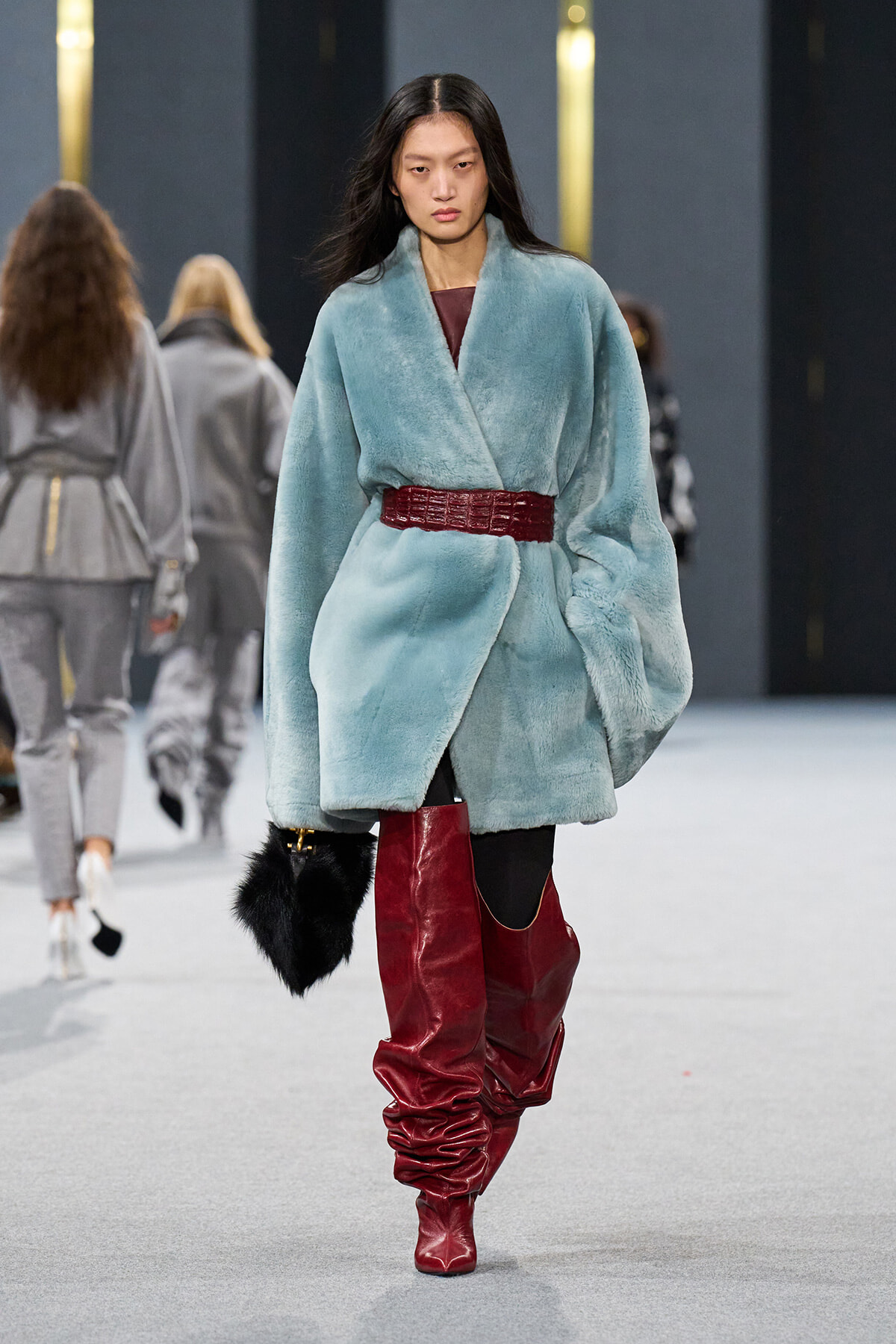 Model walking a runway in a light teal fur coat with a red belt and tall red boots, carrying a black fur handbag.