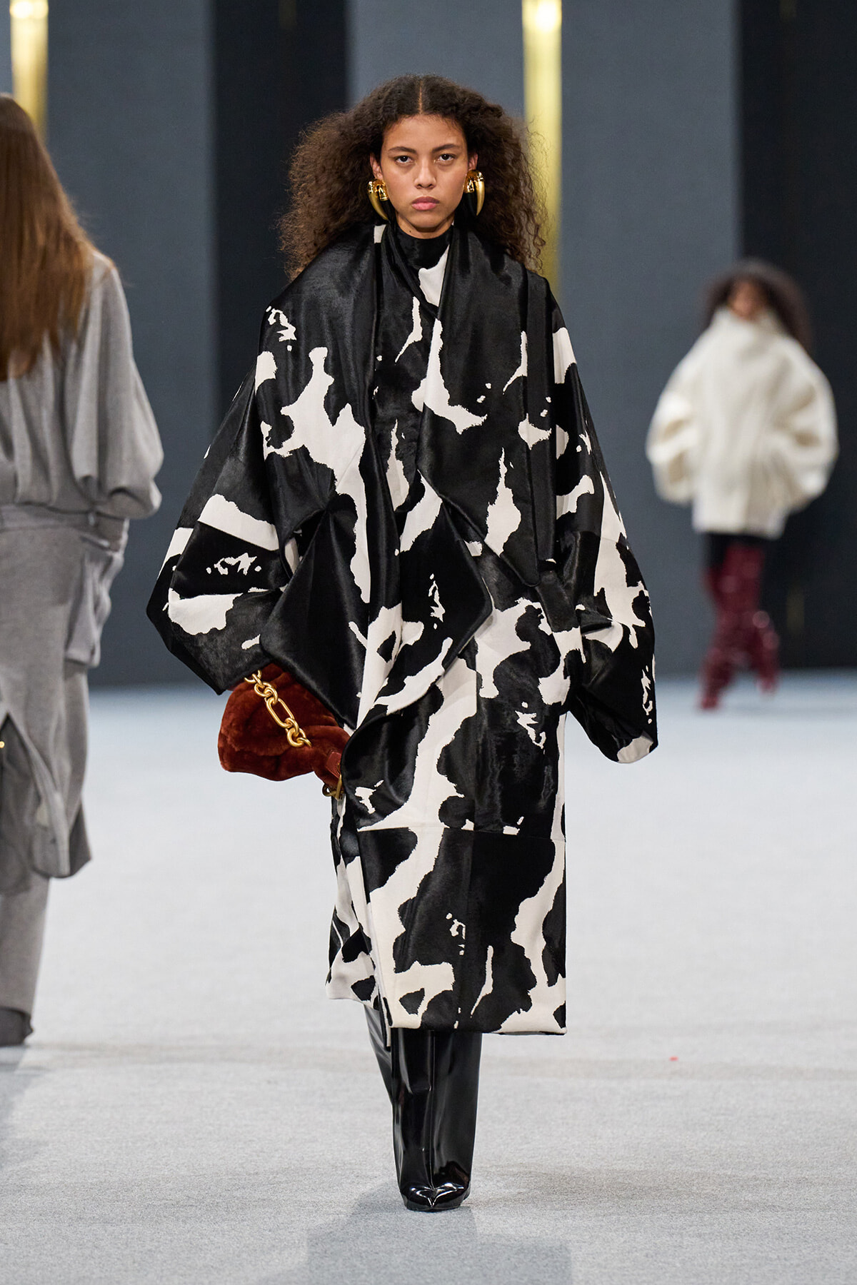 Model walking runway in an oversized black-and-white abstract coat, black patent boots, gold hoop earrings, and a brown plush handbag with a gold chain.