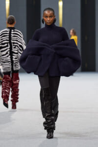 Model walking a runway in a navy oversized ribbed knit sweater with voluminous sculpted sleeves and black knee-high boots.