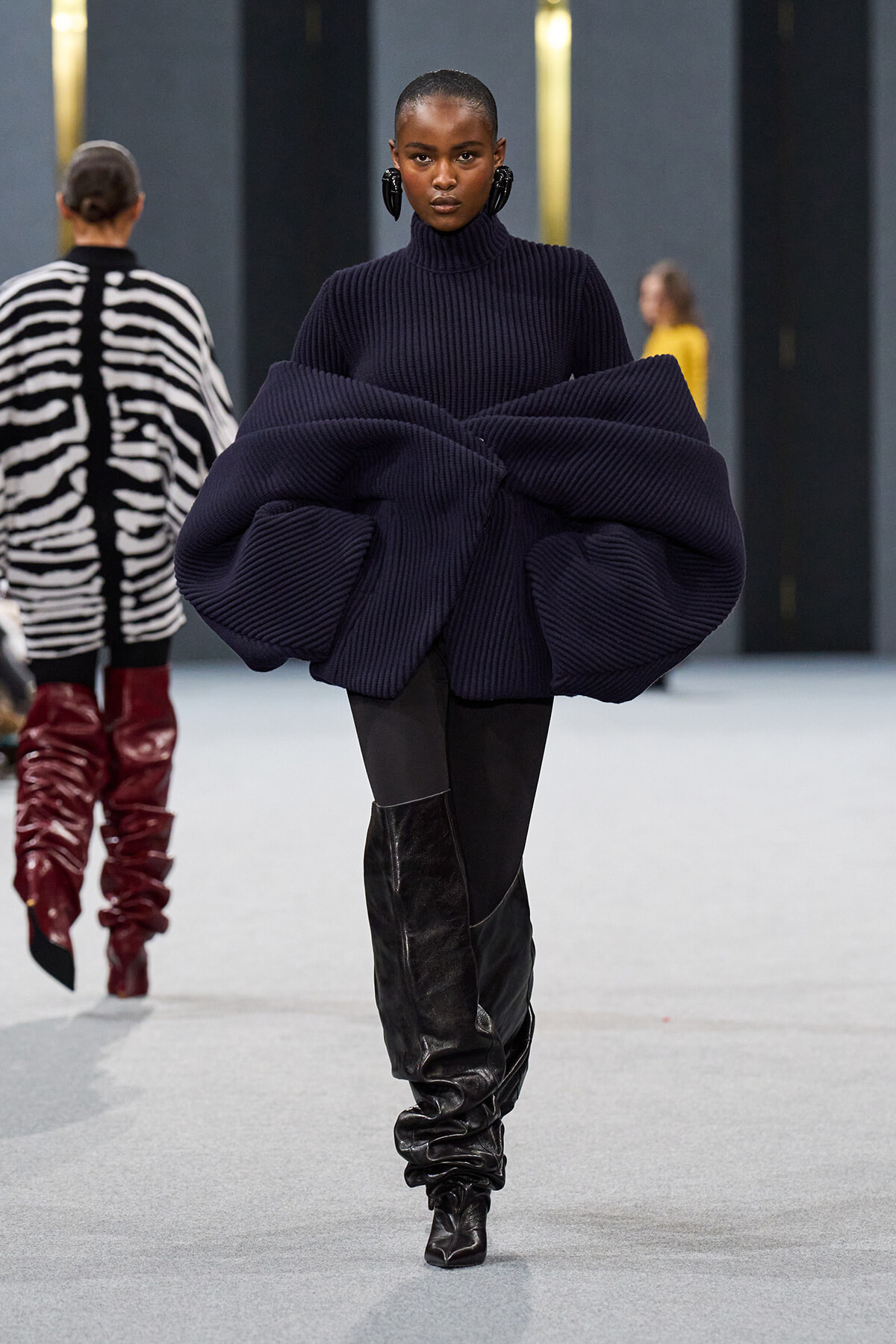 Model walking a runway in a navy oversized ribbed knit sweater with voluminous sculpted sleeves and black knee-high boots.
