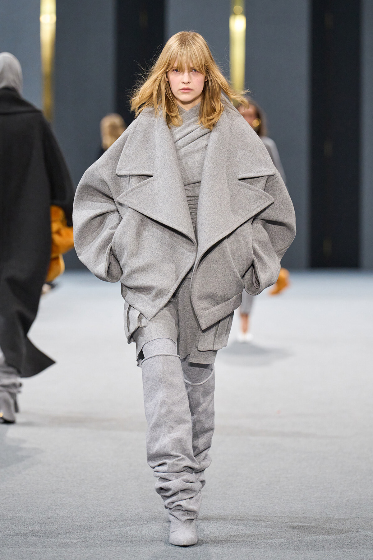 Model walks the runway in a voluminous gray wrap coat with oversized folds and draped panels.