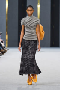 Model walking the runway in a gray draped top and a black woven skirt, paired with yellow shoes.