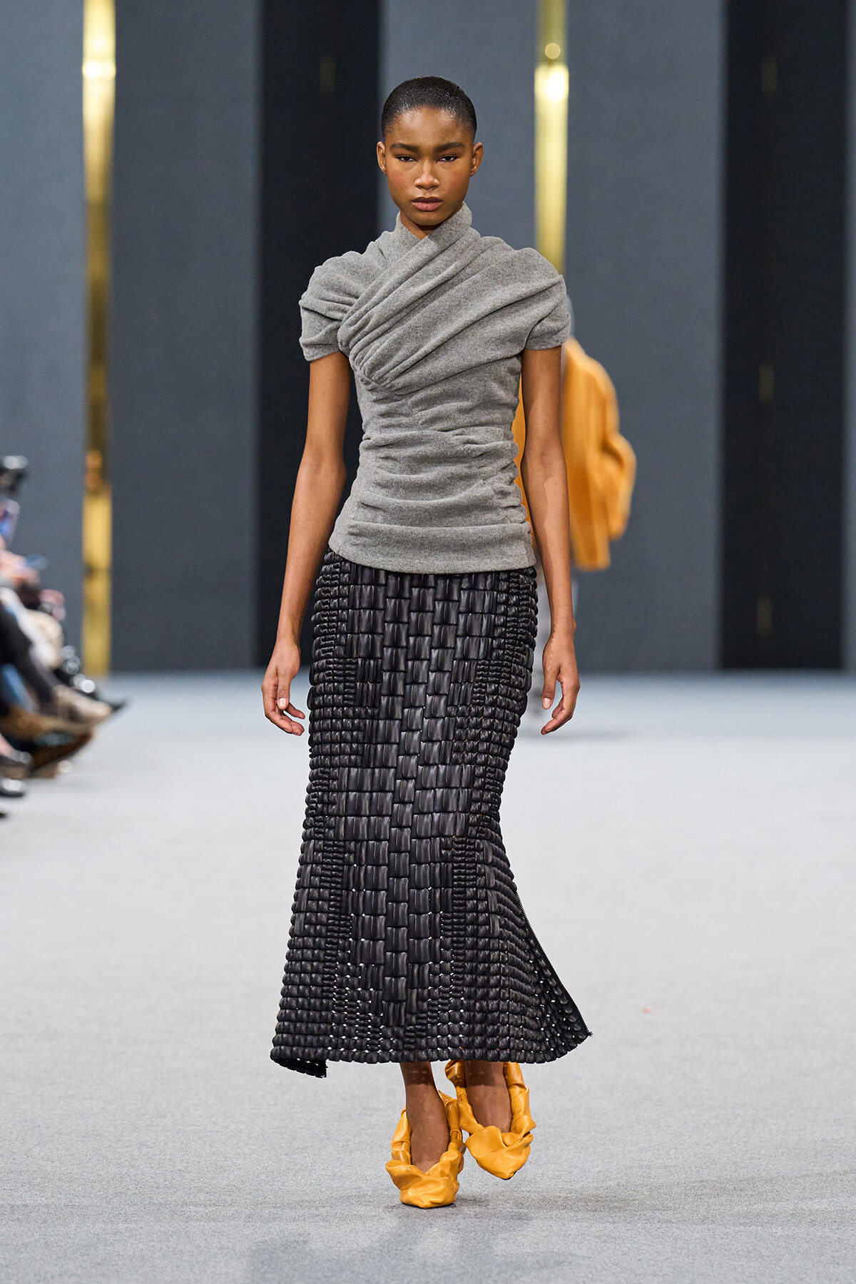Model walking the runway in a gray draped top and a black woven skirt, paired with yellow shoes.