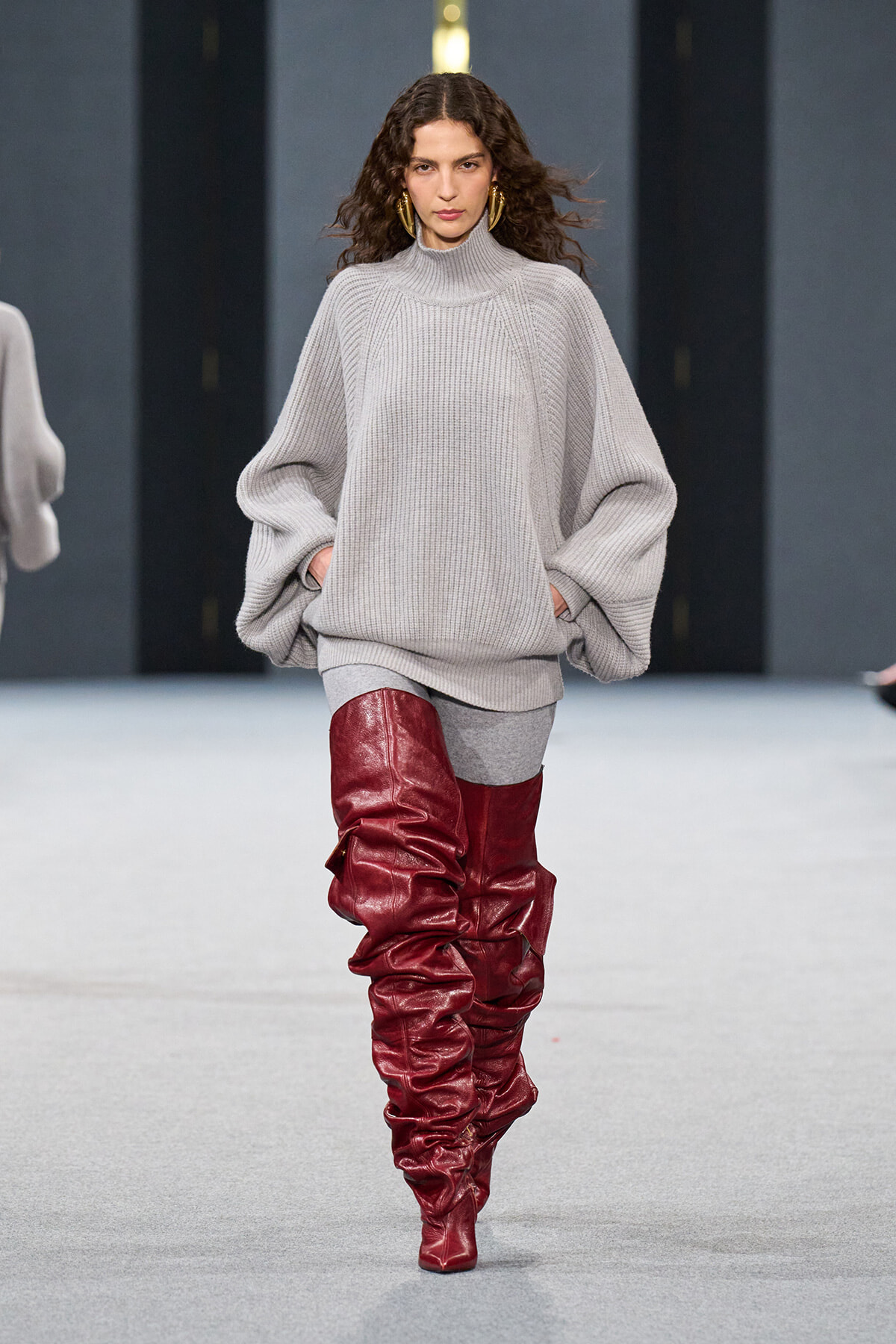 Model walking a fashion runway in an oversized light gray knit turtleneck sweater and tall red slouch boots, confident pose with hands in pockets.