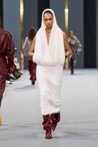 Model walks the runway in a white knitted sleeveless hooded dress with a draped front, paired with burgundy knee-high boots on a fashion show catwalk.