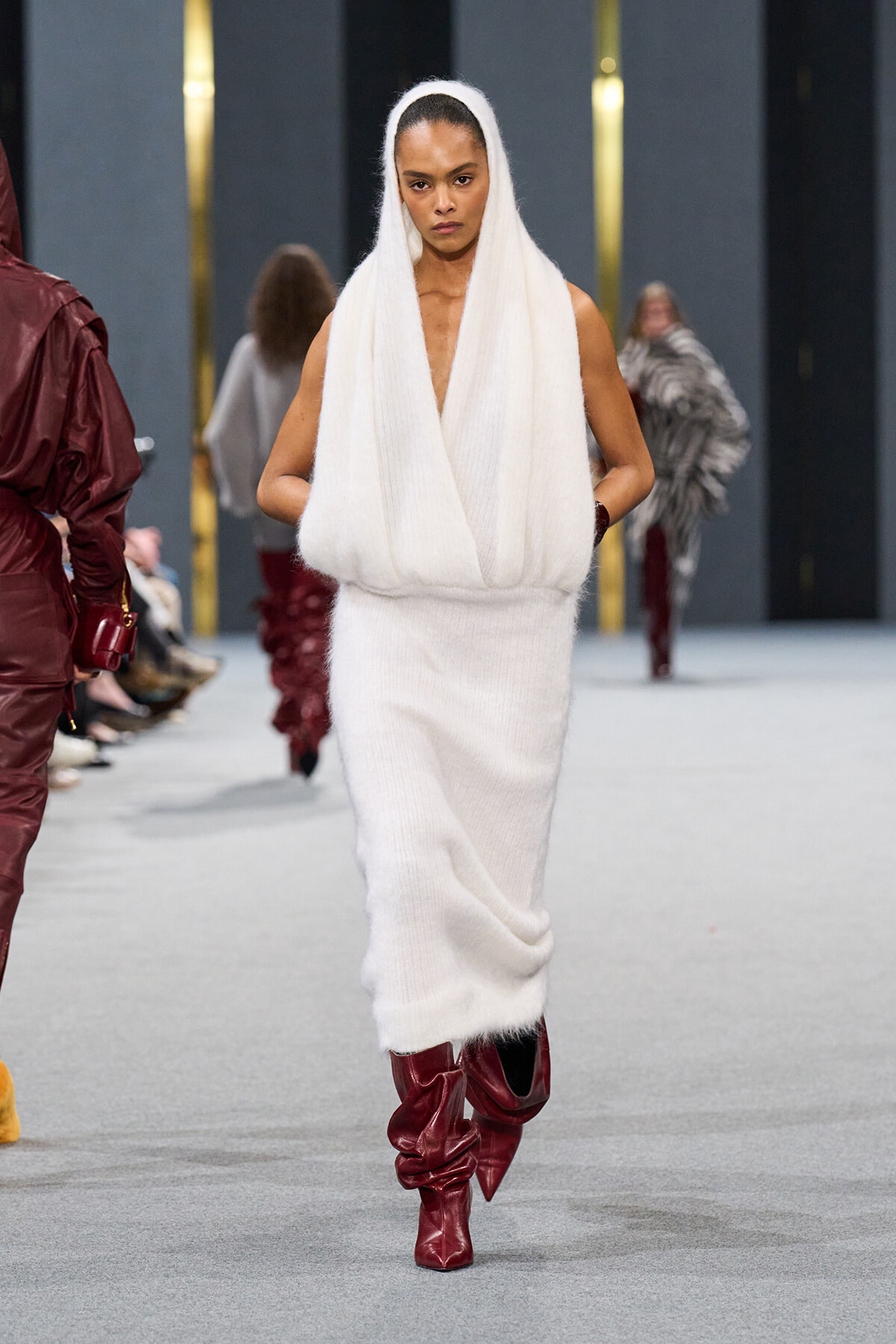 Model walks the runway in a white knitted sleeveless hooded dress with a draped front, paired with burgundy knee-high boots on a fashion show catwalk.