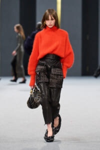 Model on a runway wearing a bright orange oversized turtleneck sweater with black leather cargo shorts, carrying a round striped handbag and wearing black shoes.