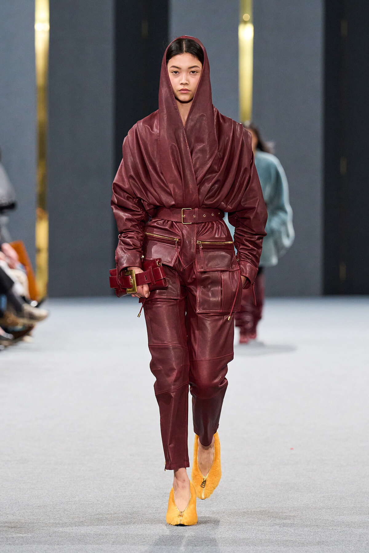 Model walking a fashion runway in a burgundy leather hooded jumpsuit with a belt and yellow suede heels, pockets visible on the outfit.