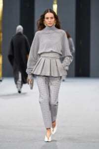 Model on a fashion runway wearing a light gray turtleneck sweater with a structured peplum and matching textured gray pants, gold earrings.