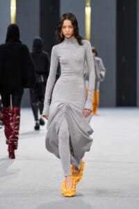 Model walks the runway in a light gray turtleneck knit dress with a dramatic gathered drape at the skirt and bright yellow shoes.