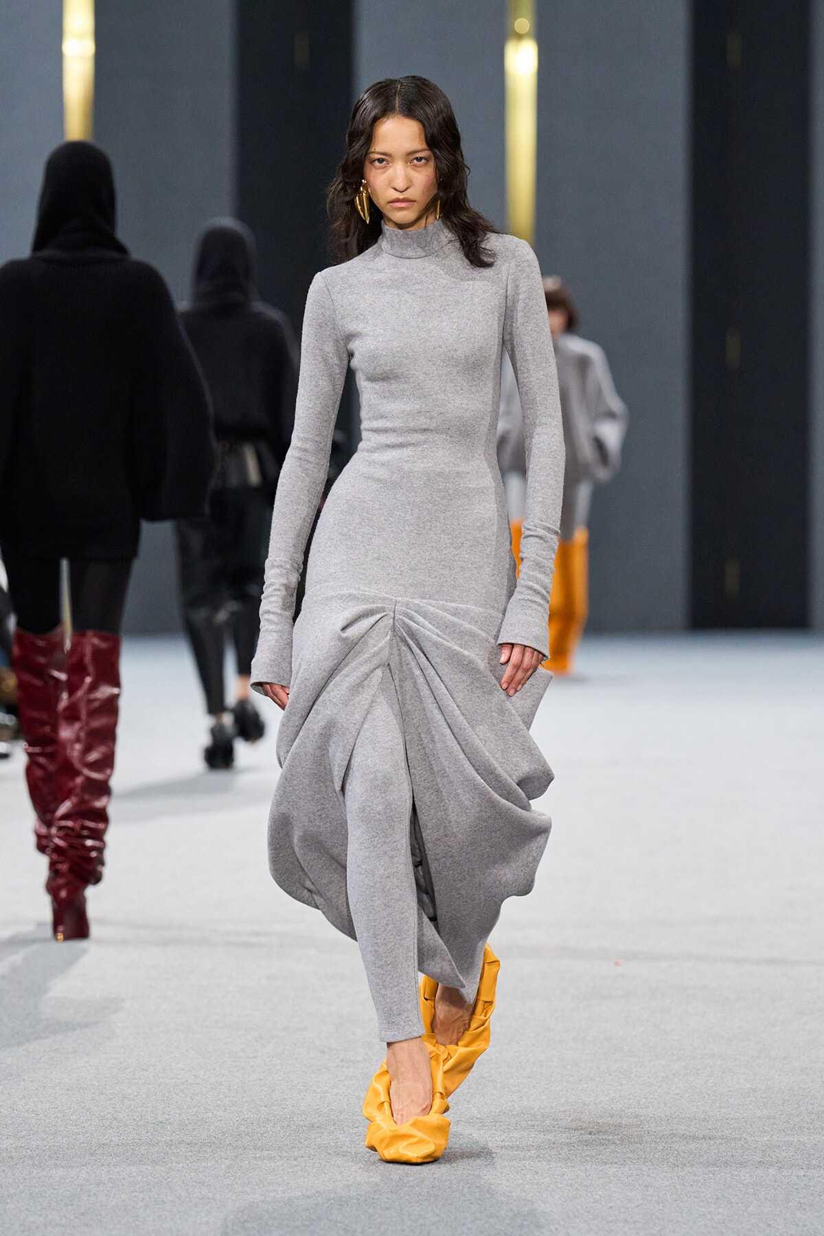 Model walks the runway in a light gray turtleneck knit dress with a dramatic gathered drape at the skirt and bright yellow shoes.