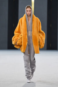 Model on runway wearing oversized orange faux-fur coat over a gray hooded jumpsuit with tall gray slouch boots.