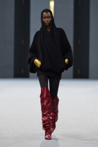 Model on a runway in an oversized black knit sweater with hood, black leggings, and tall red slouch boots accented by gold cuffs