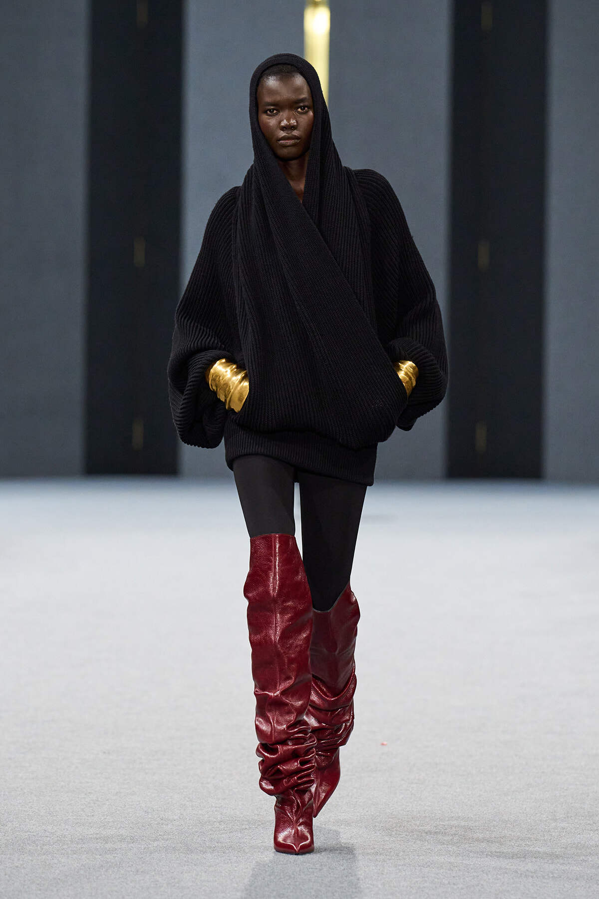 Model on a runway in an oversized black knit sweater with hood, black leggings, and tall red slouch boots accented by gold cuffs