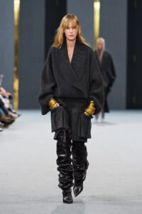 Model on a runway wearing an oversized dark knit coat with voluminous sleeves, gold cuffs, and black knee-high boots.