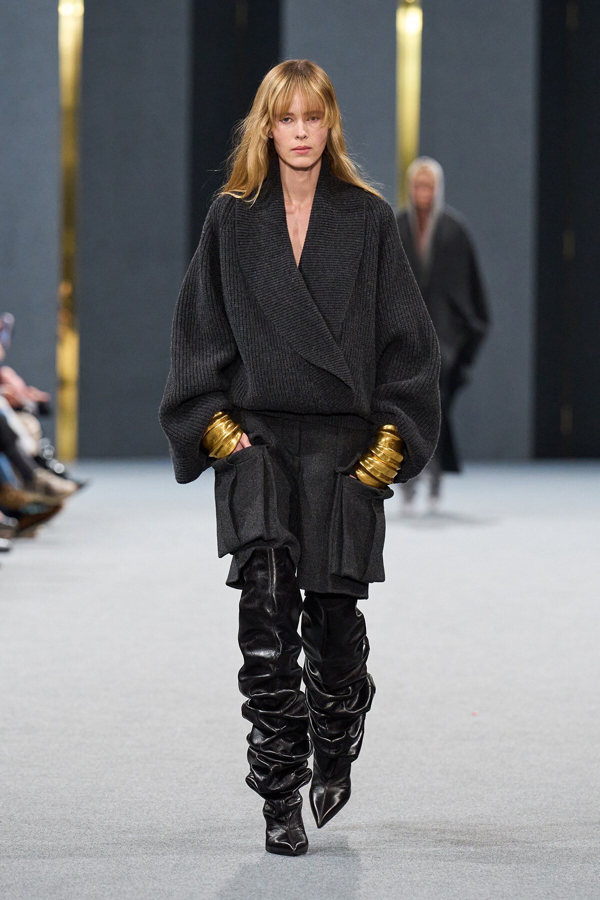Model on a runway wearing an oversized dark knit coat with voluminous sleeves, gold cuffs, and black knee-high boots.