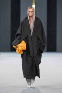 Model walks a runway in an oversized dark grey coat with a hood, holding a bright orange fur muff.