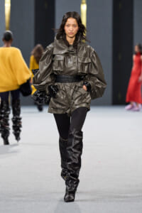 Model walking the runway in a oversized metallic gray utility jacket with large pockets, a wide belt, black gloves and knee‑high black boots.