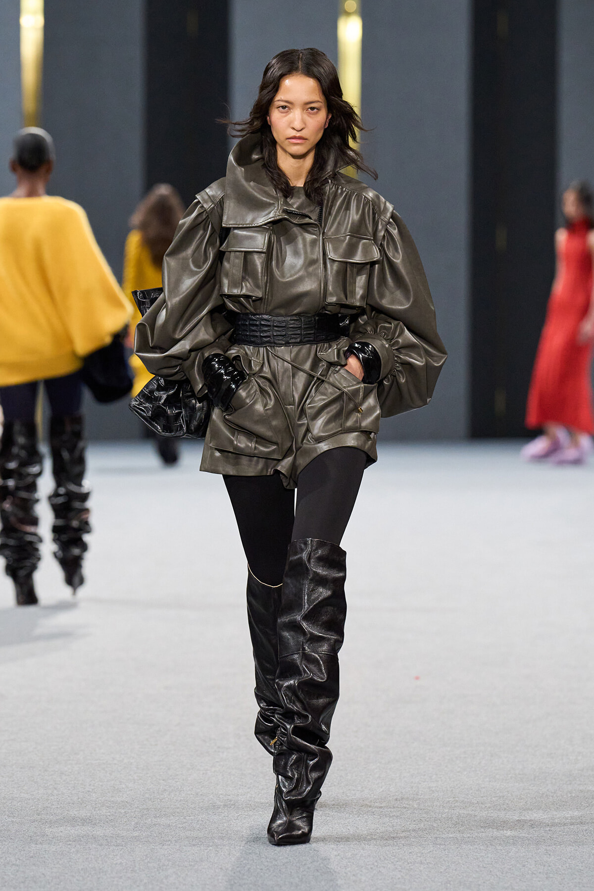 Model walking the runway in a oversized metallic gray utility jacket with large pockets, a wide belt, black gloves and knee‑high black boots.
