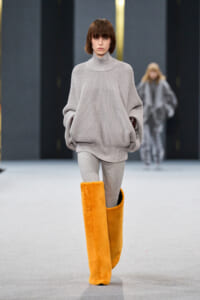 Model walks the runway in an oversized light gray turtleneck sweater with gray leggings and bright orange knee-high fuzzy boots on a fashion show catwalk.