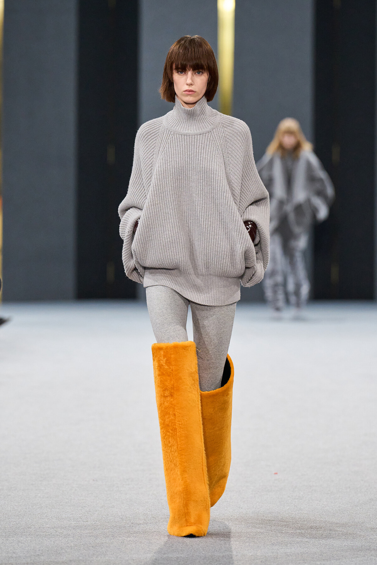 Model walks the runway in an oversized light gray turtleneck sweater with gray leggings and bright orange knee-high fuzzy boots on a fashion show catwalk.