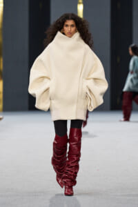 Model walking runway in an oversized cream poncho sweater with high collar, paired with black leggings and tall red ruched leather boots.