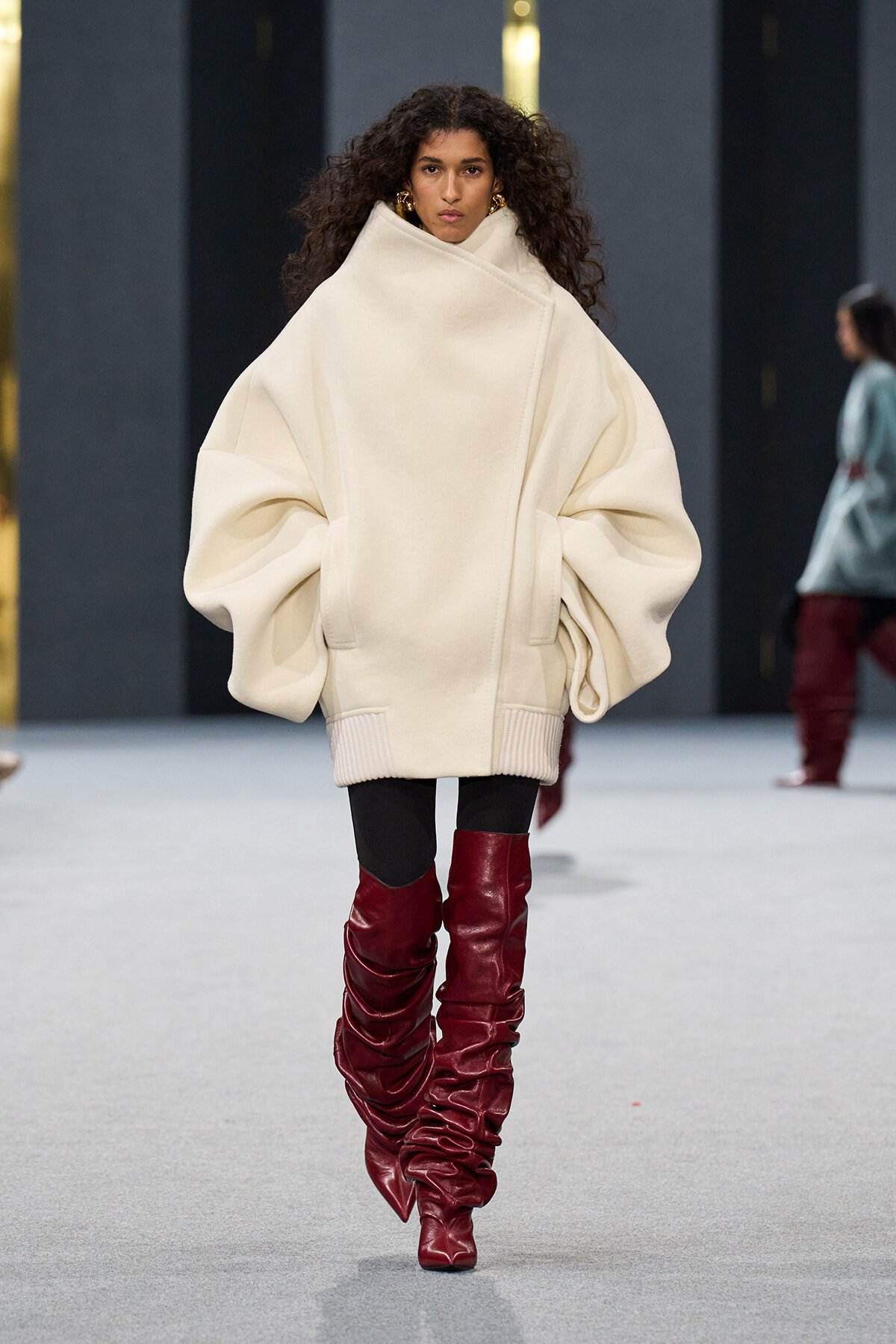 Model walking runway in an oversized cream poncho sweater with high collar, paired with black leggings and tall red ruched leather boots.