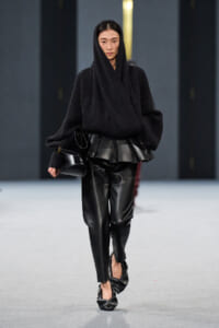 Model walking runway in an all-black ensemble: oversized ribbed sweater with hood, sculpted leather waist piece, and black leather pants, carrying a small black bag