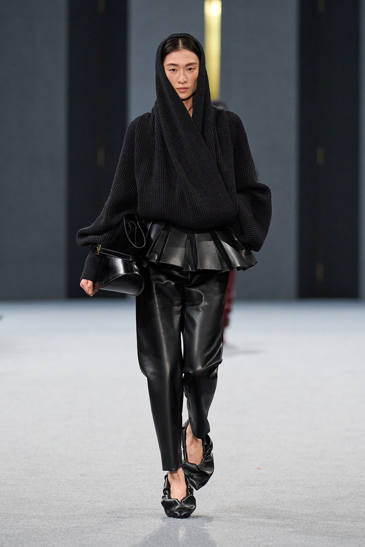 Model walking runway in an all-black ensemble: oversized ribbed sweater with hood, sculpted leather waist piece, and black leather pants, carrying a small black bag