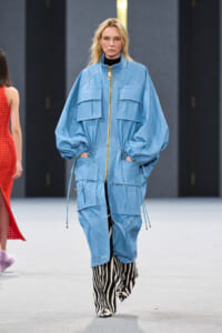 Model walks a fashion runway in a oversized light-blue utility jacket with gold zipper and large pockets, paired with zebra-print boots/pants.