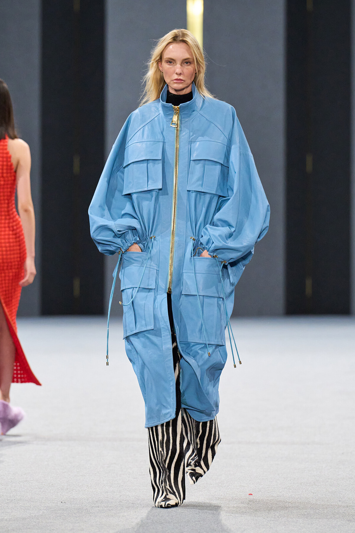 Model walks a fashion runway in a oversized light-blue utility jacket with gold zipper and large pockets, paired with zebra-print boots/pants.