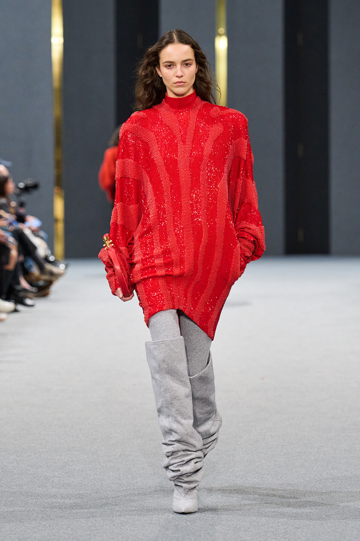 Runway model wearing an oversized red sequined sweater-dress with gray slouch boots.