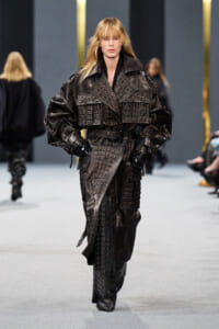 Female model on a runway wearing a large, dark crocodile-embossed leather trench with a wide belt and gloves.